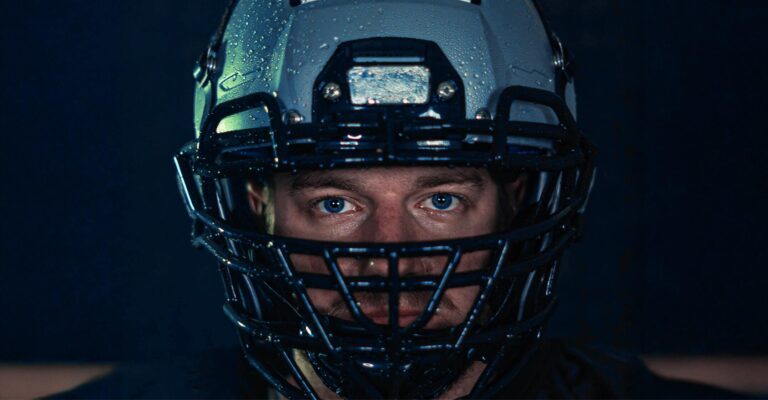 Close-up of an American football player wearing a rain-flecked helmet and facemask with intense blue eyes against a dark background.