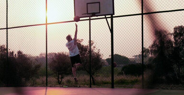 Basketball player leaping for a layup on an outdoor court behind a chain-link fence at sunset.