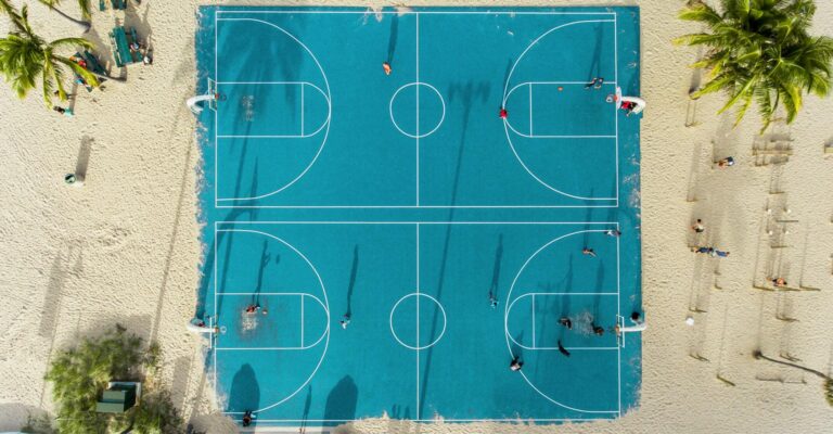 Aerial top-down view of a turquoise beachside basketball court with players, framed by sand and palm trees.