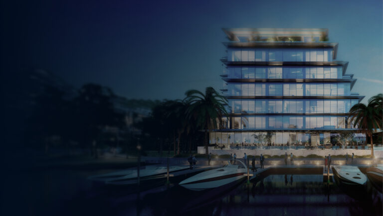 Legends One headquarters at ONE Kane on Bay Harbor Island in Miami: a modern multi-storey glass office building illuminated at dusk on the waterfront, with private yachts moored at the dock and palm trees in the foreground.