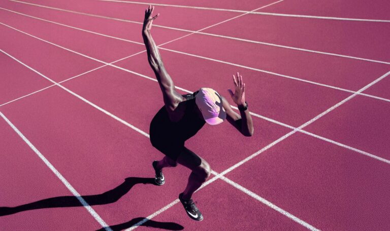 Sprinter exploding into the first stride out of the blocks on a magenta running track, conveying performance and ambition.