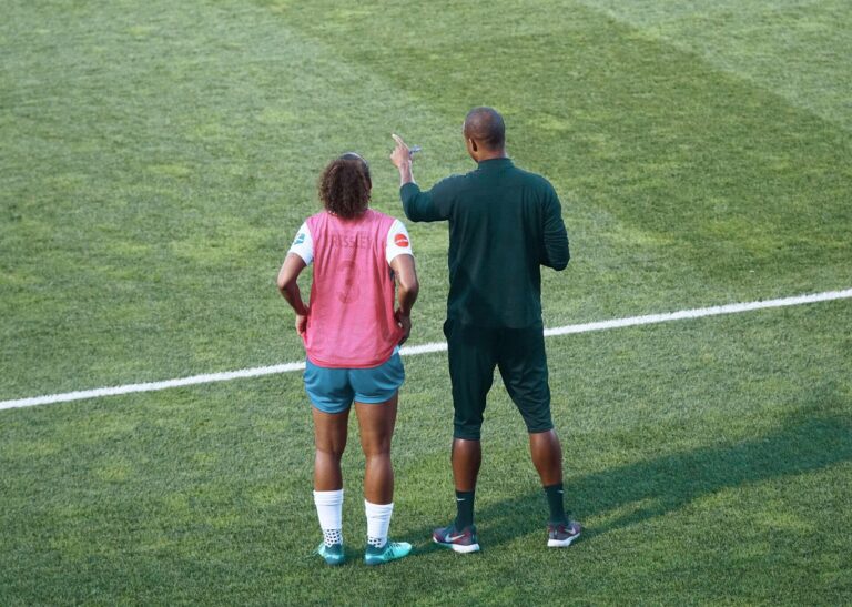 Football coach giving instructions to a player wearing a pink training bib on a grass pitch.