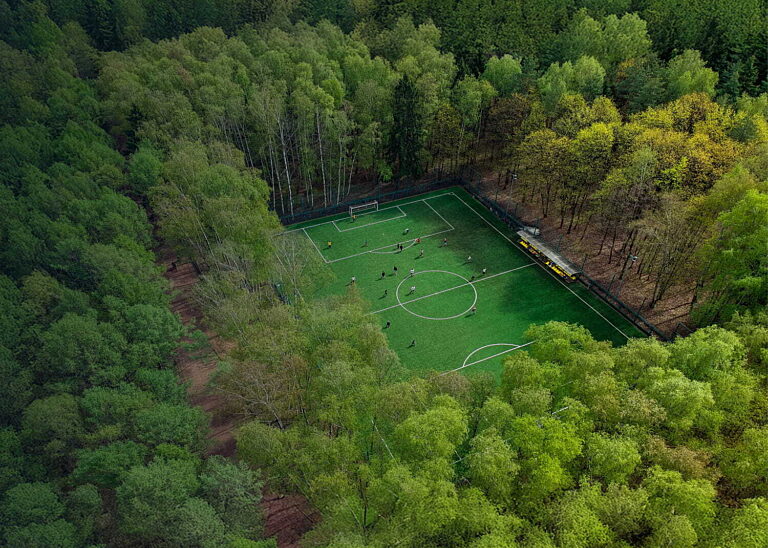 Aerial view of a small football pitch surrounded by dense green forest with players on the turf, illustrating sustainability and social responsibility in sport.