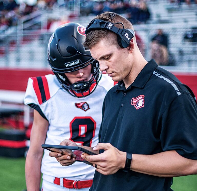 American football coach with a headset reviewing plays on a tablet beside a young helmeted player.